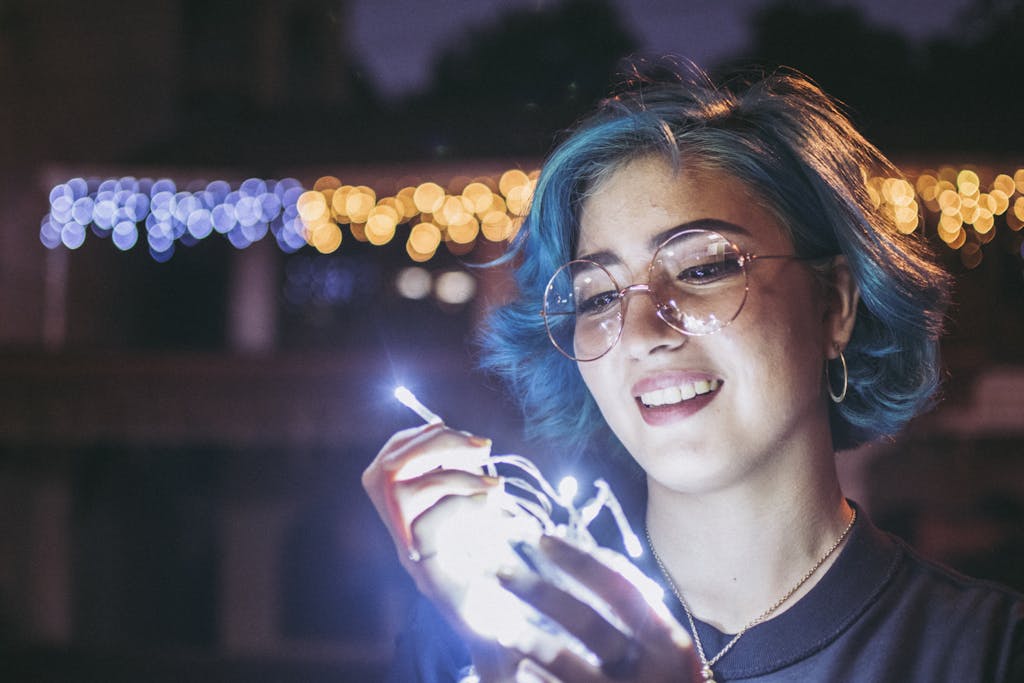 Young woman with blue hair holding string lights happily during night with colorful bokeh.